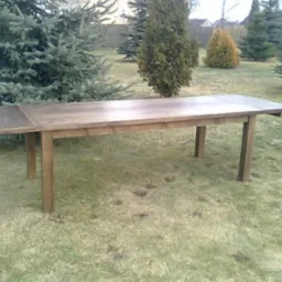 Rozkładany stół z drewna na trawie w Ziębicach, otoczony iglakami.