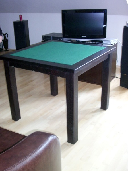 Ciemny, kwadratowy stół z zielonym blatem, ustawiony w pokoju z telewizorem i głośnikami. Widoczny fragment skórzanego fotela.