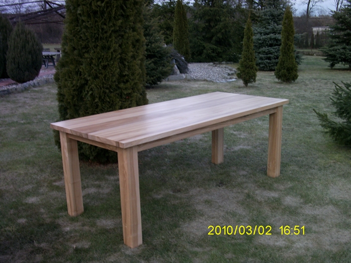 Prostokątny, jasny stół z litego drewna z grubymi nogami, ustawiony na trawniku w ogrodzie z iglakami w tle.