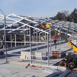 TRASCO - Rozbiórki i wyburzenia - Montaż stalowej konstrukcji hali z użyciem podnośnika teleskopowego JCB. Widoczni pracownicy na rusztowaniu i podnośniku. W tle las i budynek z cegły.