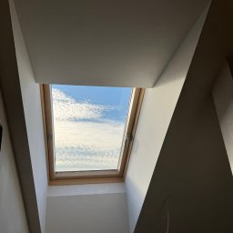 Artem Linnik - Okno dachowe z jasną drewnianą ramą, osadzone w skośnym suficie poddasza, z widokiem na błękitne niebo i puszyste chmury. Wokół gładkie, białe ściany z widocznym cieniem.