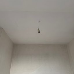Artem Linnik - Puste pomieszczenie z gładkim, jasnym sufitem i świeżo otynkowanymi ścianami w neutralnym kolorze, z pojedynczą żarówką wiszącą na kablu, gotowe do dalszych prac.
