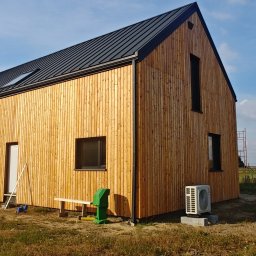 Pasyvo - Nowoczesny dom szkieletowy z drewnianą elewacją i czarnym dachem na tle błękitnego nieba. Widoczne okna, drzwi i elementy instalacji zewnętrznej.