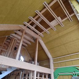 Pasyvo - Wnętrze domu szkieletowego w trakcie budowy: widoczne drewniane elementy konstrukcyjne, izolacja ścian i stropu oraz instalacja wentylacyjna z zielonych rur.