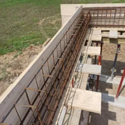 Zbrojenie w drewnianym szalunku na fundamencie budynku, widoczne podpory szalunku i teren budowy w tle.