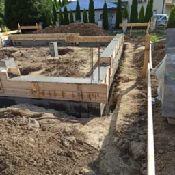 Wylane fundamenty z drewnianym szalunkiem i wystającymi prętami zbrojeniowymi, widoczne bloczki fundamentowe i wykopana ziemia, w tle dom jednorodzinny z tujami.