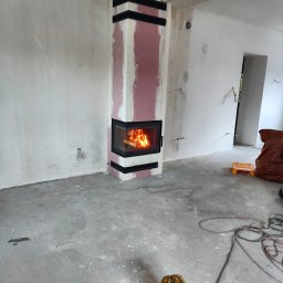 O.M KONKRET REMONTY - Narożny kominek z palącym się ogniem, wbudowany w ścianę z czarnymi listwami, w pomieszczeniu w trakcie prac wykończeniowych. Widoczne częściowo zagruntowane ściany i betonowa podłoga z kablami.