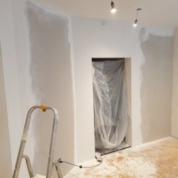 Michał Dyrduła - Puste pomieszczenie z częściowo przygotowanymi ścianami, aluminiowa drabina, otwór drzwiowy zabezpieczony folią i dwie wiszące żarówki. Na podłodze widoczny pył.