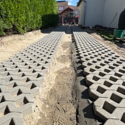 Bartosz - Dwa pasy betonowych kratek trawnikowych układane na podjeździe, z widocznym podłożem z piasku i świeżej zaprawy cementowej, w słoneczny dzień.
