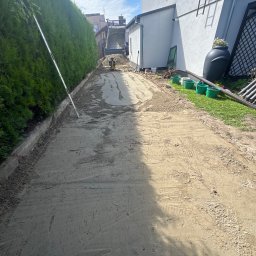 Bartosz - Długa, wąska droga gruntowa z wyrównanym podłożem, sznurkiem i poziomicą, w tle wywrotka i mała maszyna budowlana. Obok zielony żywopłot i biały budynek.
