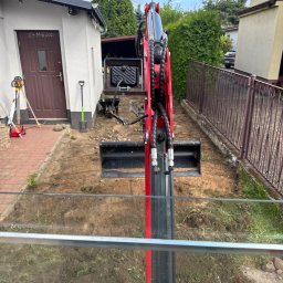 Bartosz - Widok z kabiny na ramię czerwonej minikoparki z łyżką, kopiącej ziemię obok domu. W tle laserowy niwelator i przygotowany teren pod instalacje.