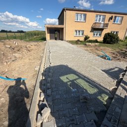 Bartosz - Układanie szarej kostki brukowej na podjeździe przed domem jednorodzinnym, widoczny krawężnik, stos ziemi i niebieska rura instalacyjna. Praca w toku, w tle pole uprawne.
