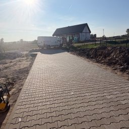 Bartosz - Świeżo ułożona ścieżka z szarej kostki brukowej, z zagęszczarką w pobliżu. W tle budynek mieszkalny, biała furgonetka i minikoparka na placu budowy w słoneczny dzień.