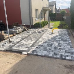 Bartosz - Podjazd z szarej kostki brukowej w pasy, częściowo ułożony przed domem. Obok stosy materiału, maszyna budowlana i żółta rura instalacyjna. W tle dom i trawnik.