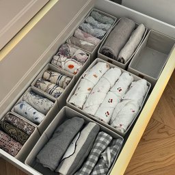 Posprzątane Kąty by Patrycja Kamrowska - Otwarta szuflada z beżowymi organizerami, w których starannie ułożono złożoną bieliznę i ubrania w pastelowych kolorach, z koronkami, kwiatowymi i kratkowanymi wzorami, oświetlona ciepłym światłem.
