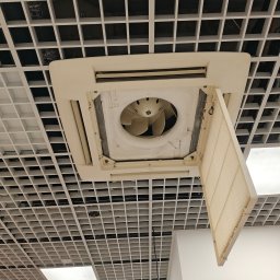 LokalnaEkipa - Zabrudzony, podwieszany klimatyzator kasetowy z otwartą klapą filtra i widocznym wentylatorem, osadzony w białym suficie podwieszanym typu grid, sugerujący potrzebę konserwacji.