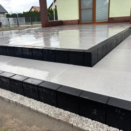 KAROL KORZENIEWSKI - Nowoczesny taras z dużych, jasnoszarych płyt i czarnych, geometrycznych stopni, lśniący po deszczu, przy jasnym budynku.