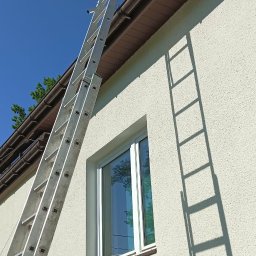 ALP Usługi Wysokościowe Marcin Adasiewicz - Długa, aluminiowa drabina z górnym stabilizatorem oparta o beżową elewację domu, obok brązowej rynny, pod bezchmurnym niebem. Widoczny cień drabiny.