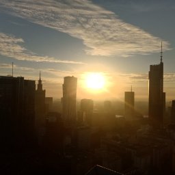 ALP Usługi Wysokościowe Marcin Adasiewicz - Wysoka panorama miejska o wschodzie słońca, z intensywnym słońcem w centrum, rzucającym złocistą mgiełkę na horyzont. Sylwetki nowoczesnych wieżowców dominują na tle nieba z rozproszonymi chmurami.