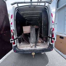 Wiktor Janiak - Biały bus dostawczy z szeroko otwartymi tylnymi drzwiami, ukazujący wnętrze wypełnione kartonami, panelami i drobnymi odpadami. Widoczny bagażnik dachowy.