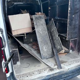 Wiktor Janiak - Otwarty tył białego busa dostawczego z brudną podłogą, wypełniony kartonami, ciemnymi płytami i drewnianymi elementami; widoczny żółty hak holowniczy.