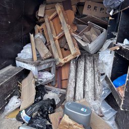 Wiktor Janiak - Wnętrze busa wypełnione różnorodnymi odpadami: starymi drewnianymi ramami, kartonami, czarnymi workami na śmieci, plastikowymi opakowaniami i zniszczonym drewnianym stołkiem.