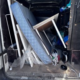 Wiktor Janiak - Tył białego busa załadowany niebieskim materacem, białym stelażem łóżka, jasnym stołem i różnymi rzeczami, sugerującymi wywózkę lub przewóz.