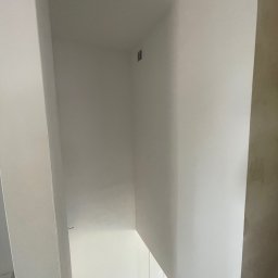 Łukasz Len - Gładkie, jednolicie pomalowane ściany i sufit w jasnej klatce schodowej, ujęcie z góry, z widocznym drewnianym parkietem i drobnym otworem wentylacyjnym.