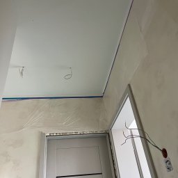 Łukasz Len - Wnętrze pomieszczenia w trakcie prac wykończeniowych: świeżo pomalowany sufit, ściany zabezpieczone folią i niebieską taśmą malarską, obok nowoczesne szare drzwi i odkryte okablowanie elektryczne.