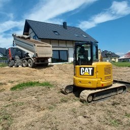 Matkop - Żółta minikoparka CAT i wywrotka z podniesioną skrzynią na placu budowy w Golkowicach, gdzie przygotowywany jest grunt przy nowoczesnym domu z ciemnym dachem, pod błękitnym niebem.
