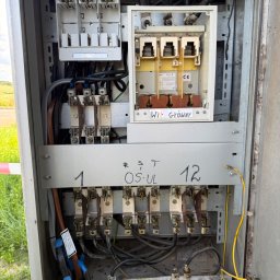 Michał Pawłowski - Otwarta szafa elektryczna z bezpiecznikami, grubymi kablami, głównym wyłącznikiem i oznaczeniami R S T; na dole luźne wkładki topikowe, w tle zielone pole.
