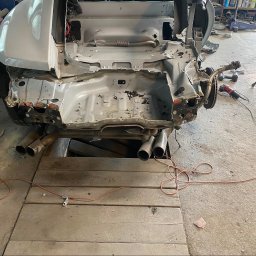 CarSow blacharstwo lakiernictwo pojazdowe - Srebrne auto w Częstochowie z demontażem tylnej części karoserii, ukazującym podwozie i rury wydechowe nad kanałem serwisowym. Na podłodze szlifierka kątowa.
