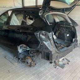 CarSow blacharstwo lakiernictwo pojazdowe - Czarne auto z otwartym bagażnikiem i zdemontowanym tyłem w warsztacie, widoczne okablowanie, zawieszenie, brak koła i narzędzia na ceglanej posadzce.
