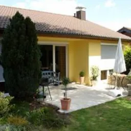 Jasnożółty dom jednorodzinny z brązowym dachem i betonowym tarasem, widok z zadbanego ogrodu.