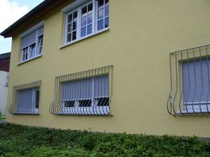 Żółta elewacja dwupiętrowego budynku z oknami zabezpieczonymi metalowymi kratami i częściowo zasłoniętymi roletami zewnętrznymi.