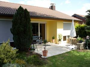 Jasnożółty dom jednorodzinny z brązowym dachem i betonowym tarasem, widok z zadbanego ogrodu.