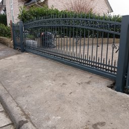 MPN Zaune - Ciemnoszara, kuta brama wjazdowa przesuwna z ozdobnymi spiralami i szpiczastymi zakończeniami, wyposażona w lampę ostrzegawczą na słupku, na tle betonowego podjazdu.