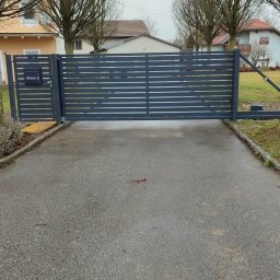 MPN Zaune - Nowoczesna, antracytowa brama przesuwna z szerokimi poziomymi lamelami, zintegrowana z furtką ze skrzynką pocztową, na tle podjazdu i domów jednorodzinnych.