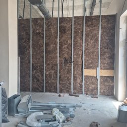 Ben-Bud Krzysztof Chrościelewski - Montaż izolacji z wełny mineralnej w metalowym stelażu ściany działowej. Widoczne elementy wentylacji pod sufitem i profile na podłodze. Etap prac wykończeniowych.