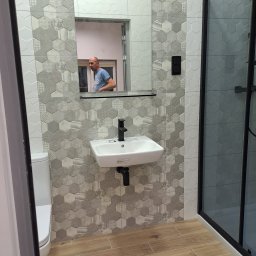 Ben-Bud Krzysztof Chrościelewski - Wykończona łazienka z heksagonalnymi płytkami w odcieniach szarości, drewnopodobną podłogą i kabiną prysznicową z czarnymi ramami. W lustrze odbija się osoba.