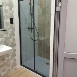 Ben-Bud Krzysztof Chrościelewski - Nowoczesna kabina prysznicowa z czarną ramą i heksagonalnymi płytkami w Białymstoku. Widoczna armatura i drewnopodobna podłoga. Wykończenie łazienki.