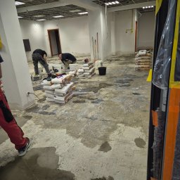 Ben-Bud Krzysztof Chrościelewski - Przygotowanie podłoża pod wylewkę w biurze: pracownicy wyrównują podłoże, widoczne worki z zaprawą, narzędzia i surowy stan wykończenia wnętrza.