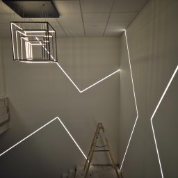 Ben-Bud Krzysztof Chrościelewski - Nowoczesne oświetlenie schodów z geometrycznymi liniami LED na ścianach i designerską lampą wiszącą w kształcie sześcianu. Widoczna drabina malarska.