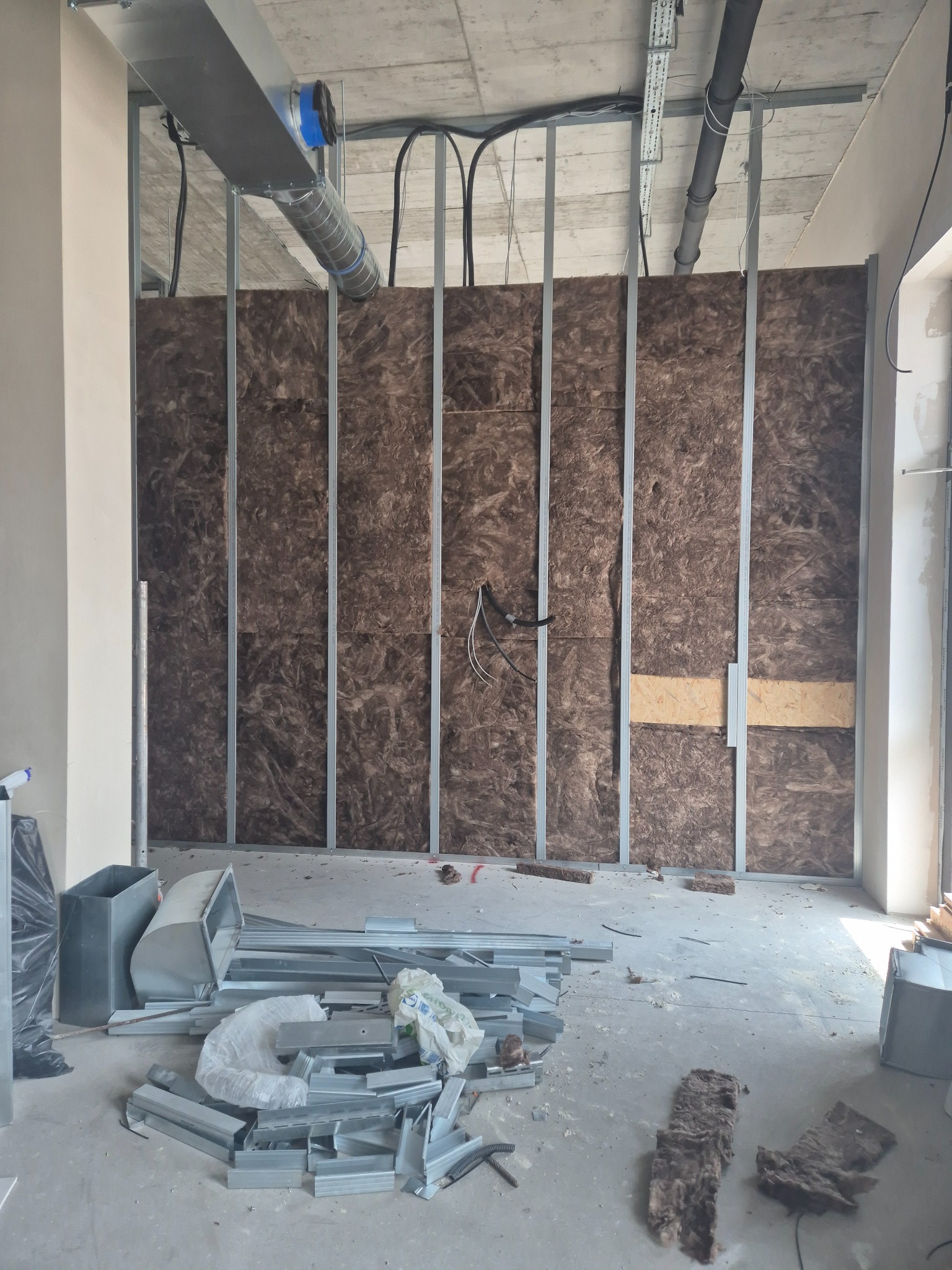 Montaż izolacji z wełny mineralnej w metalowym stelażu ściany działowej. Widoczne elementy wentylacji pod sufitem i profile na podłodze. Etap prac wykończeniowych.