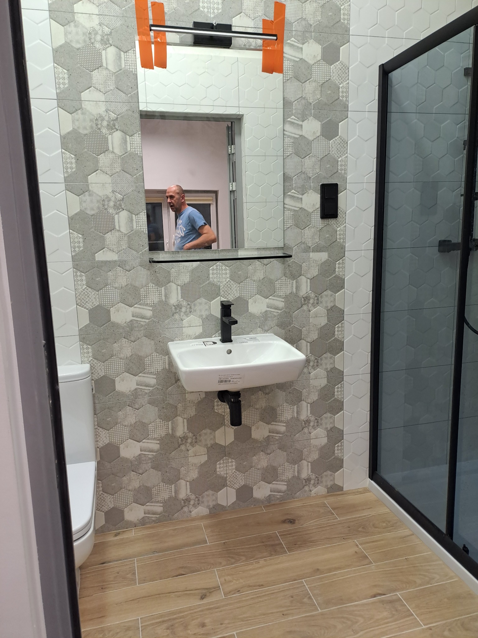 Wykończona łazienka z heksagonalnymi płytkami w odcieniach szarości, drewnopodobną podłogą i kabiną prysznicową z czarnymi ramami. W lustrze odbija się osoba.