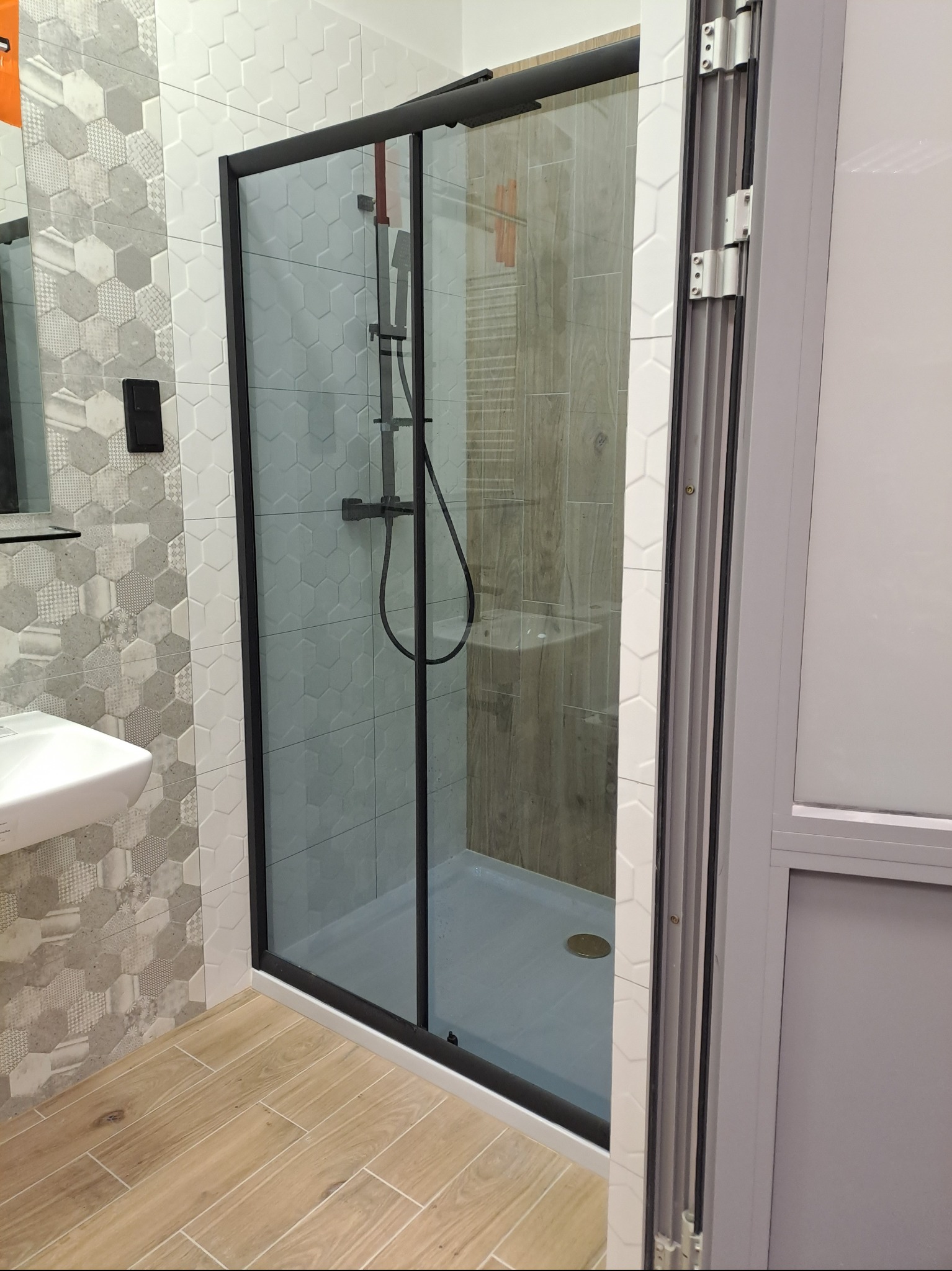 Nowoczesna kabina prysznicowa z czarną ramą i heksagonalnymi płytkami w Białymstoku. Widoczna armatura i drewnopodobna podłoga. Wykończenie łazienki.
