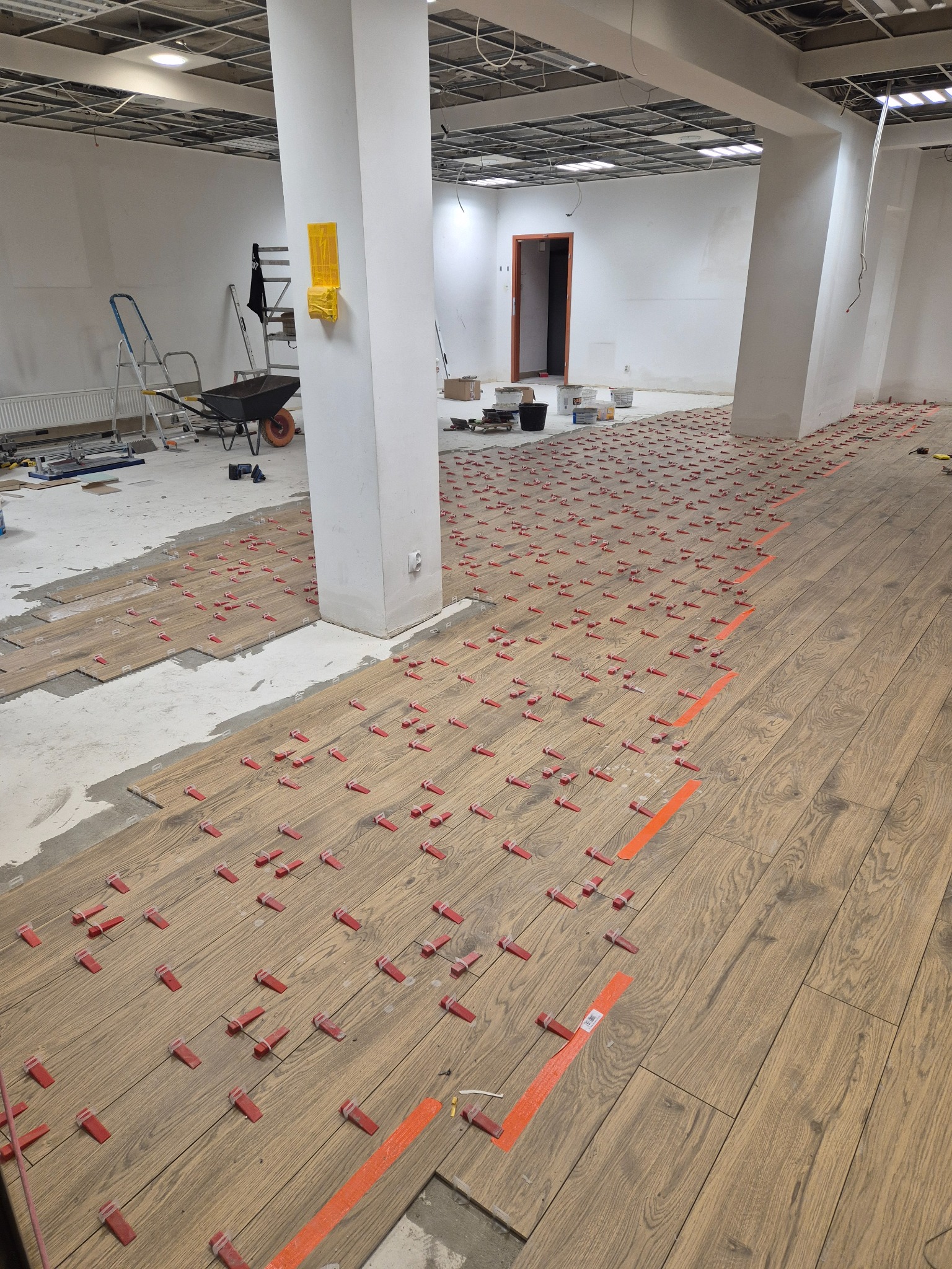 Układanie drewnopodobnych płytek podłogowych w dużym pomieszczeniu biurowym z użyciem klipsów poziomujących i taśmy wyznaczającej obszar roboczy.