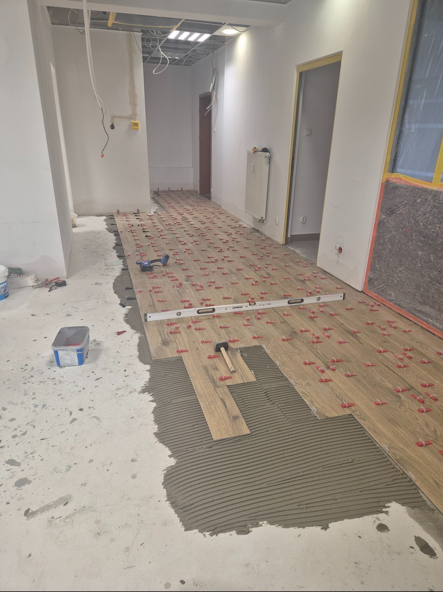 Układanie podłogi z płytek imitujących drewno w korytarzu. Widoczne poziomica, młotek i czerwone kliny dystansowe. Częściowo nałożony klej. W tle instalacje pod sufitem.