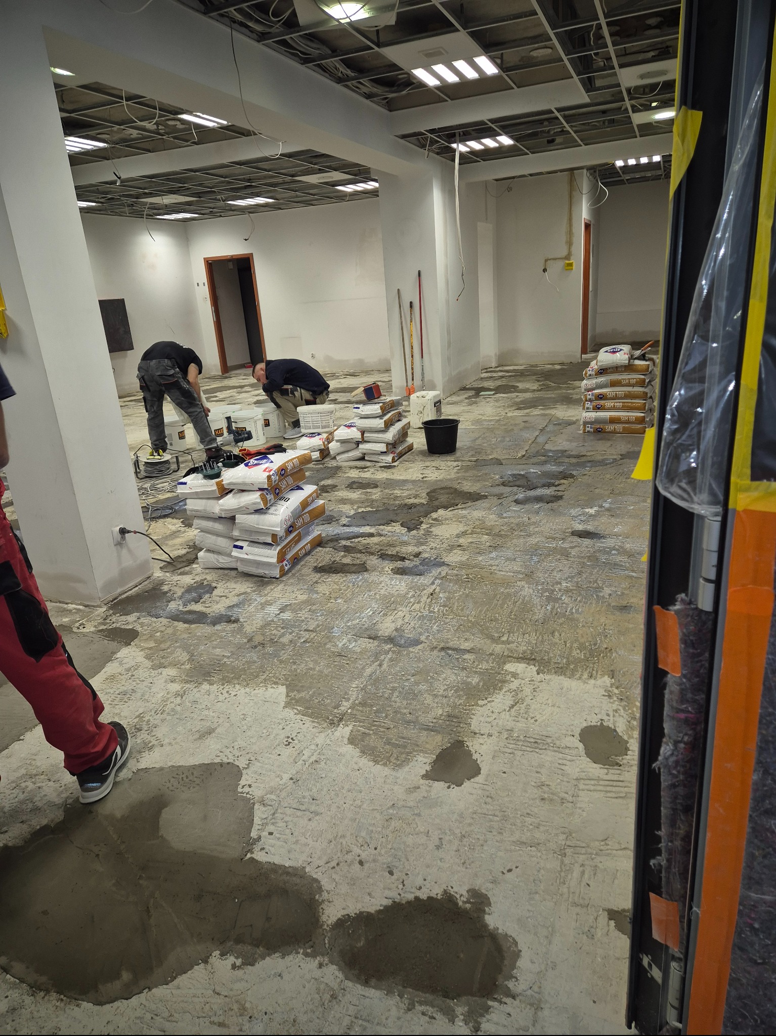 Przygotowanie podłoża pod wylewkę w biurze: pracownicy wyrównują podłoże, widoczne worki z zaprawą, narzędzia i surowy stan wykończenia wnętrza.