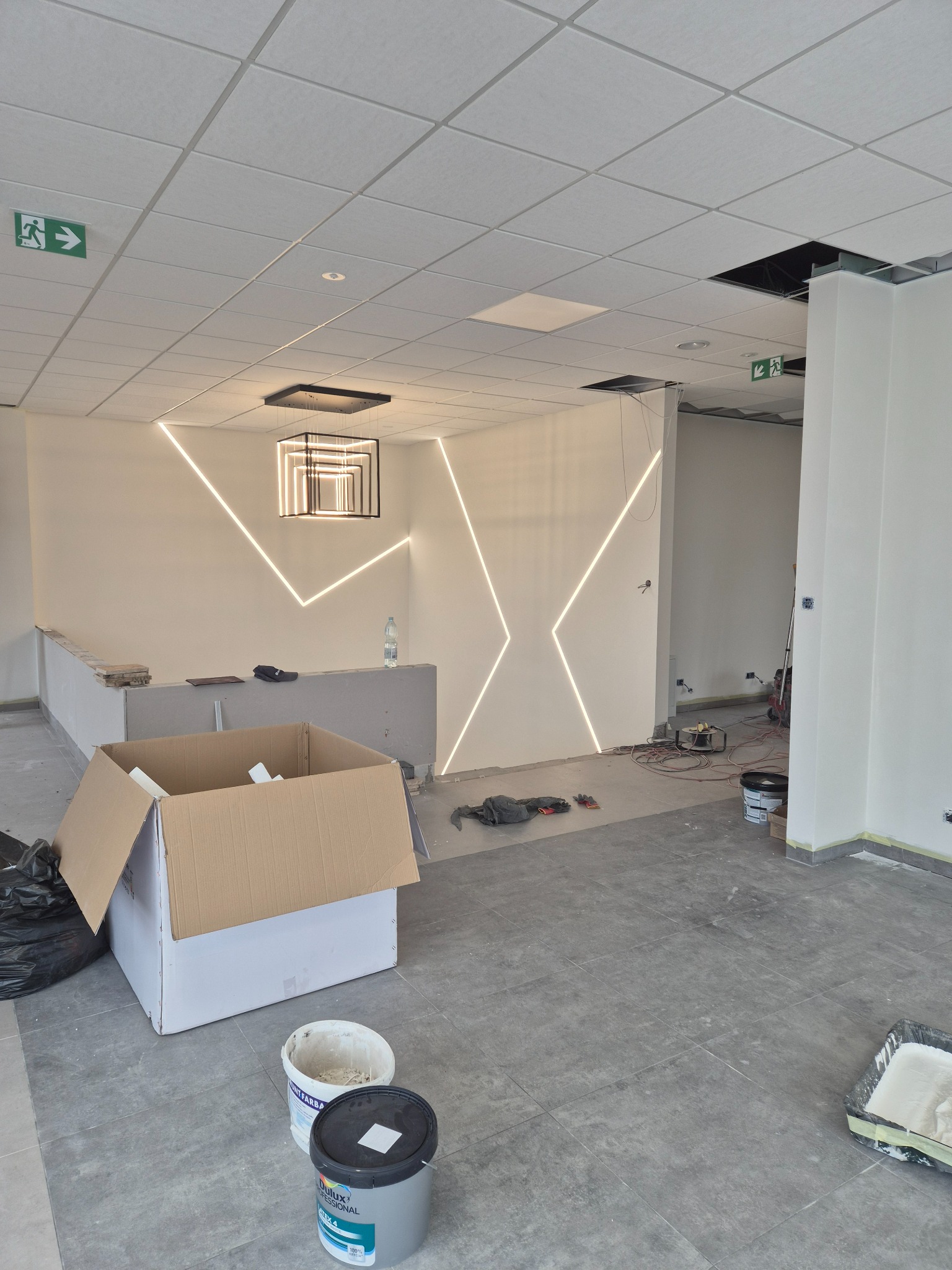 Wnętrze biura w trakcie wykańczania: sufit podwieszany, oświetlenie liniowe w kształcie litery X na ścianie, karton, puszki z farbą na szarej podłodze.